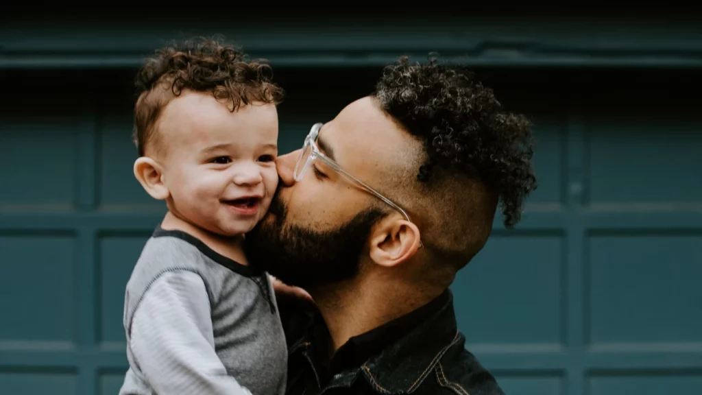 A young dad in glasses holding a little boy giving him a kiss on the cheek.